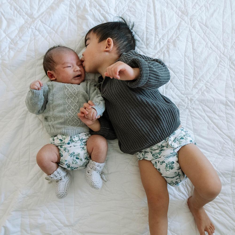 Newborn baby and toddler brother wearing snuggly knitted clothing and matching eucalyptus reusable nappies. Toddler is giving a kiss to newborn brother.