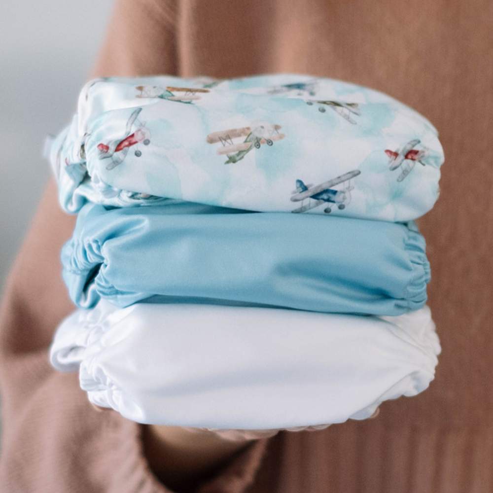 Hand of lady wearing knitted jumper holding stack of 3 cloth nappies: white, blue and blue aeroplanes.