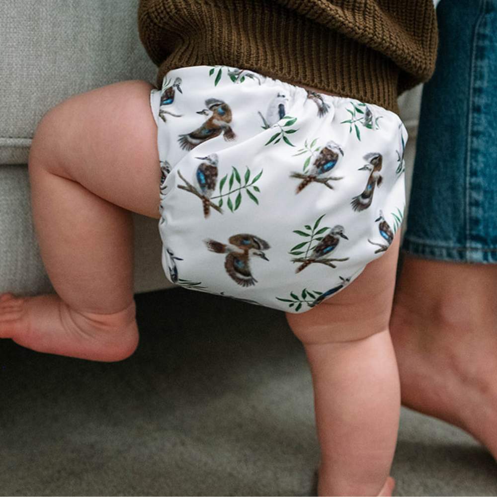 Baby standing wearing kookaburra reusable nappy lifting leg to climb onto couch with mum