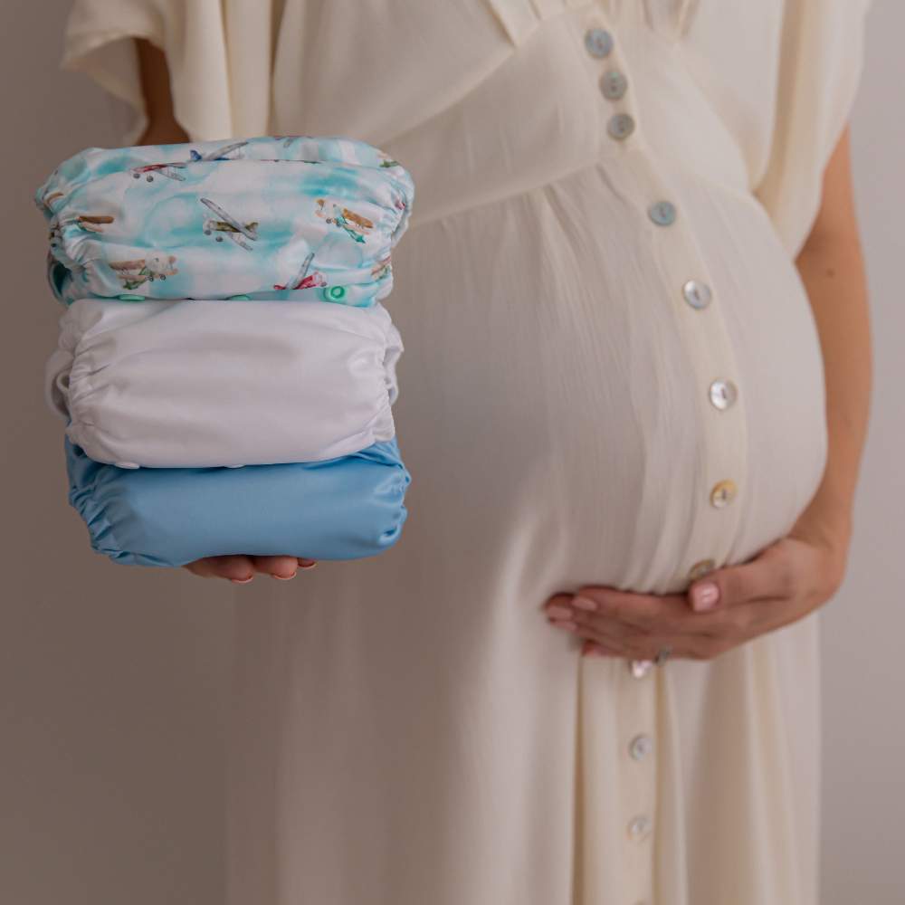 Pregnant lady holding baby bump with one hand and a stack of 3 reusable nappies in her other hand
