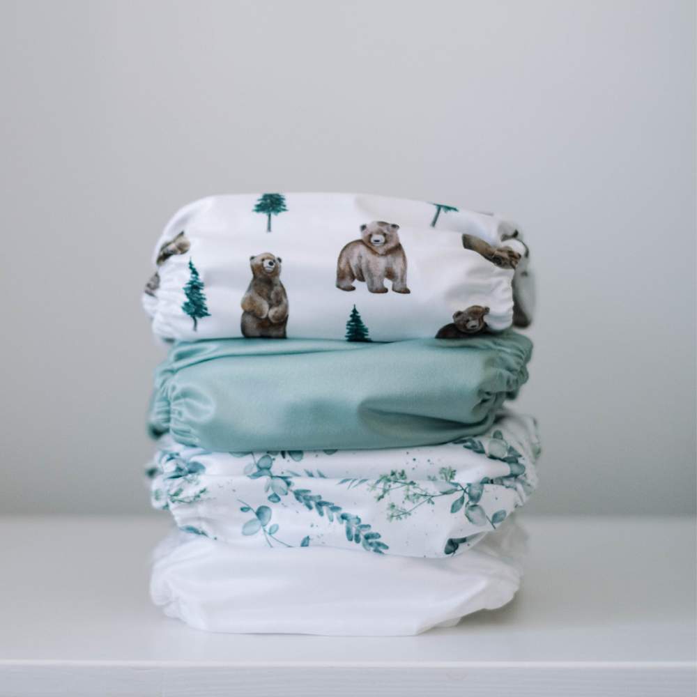 Stack of cloth diapers on white surface. Diaper prints include: white, eucalyptus, sage green and bear cubs
