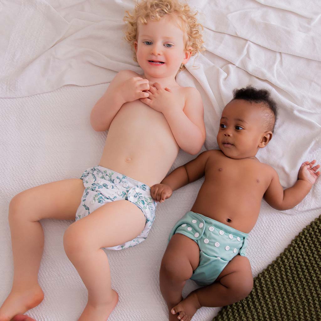 Two babies laying on a white bed wearing cloth nappies. 