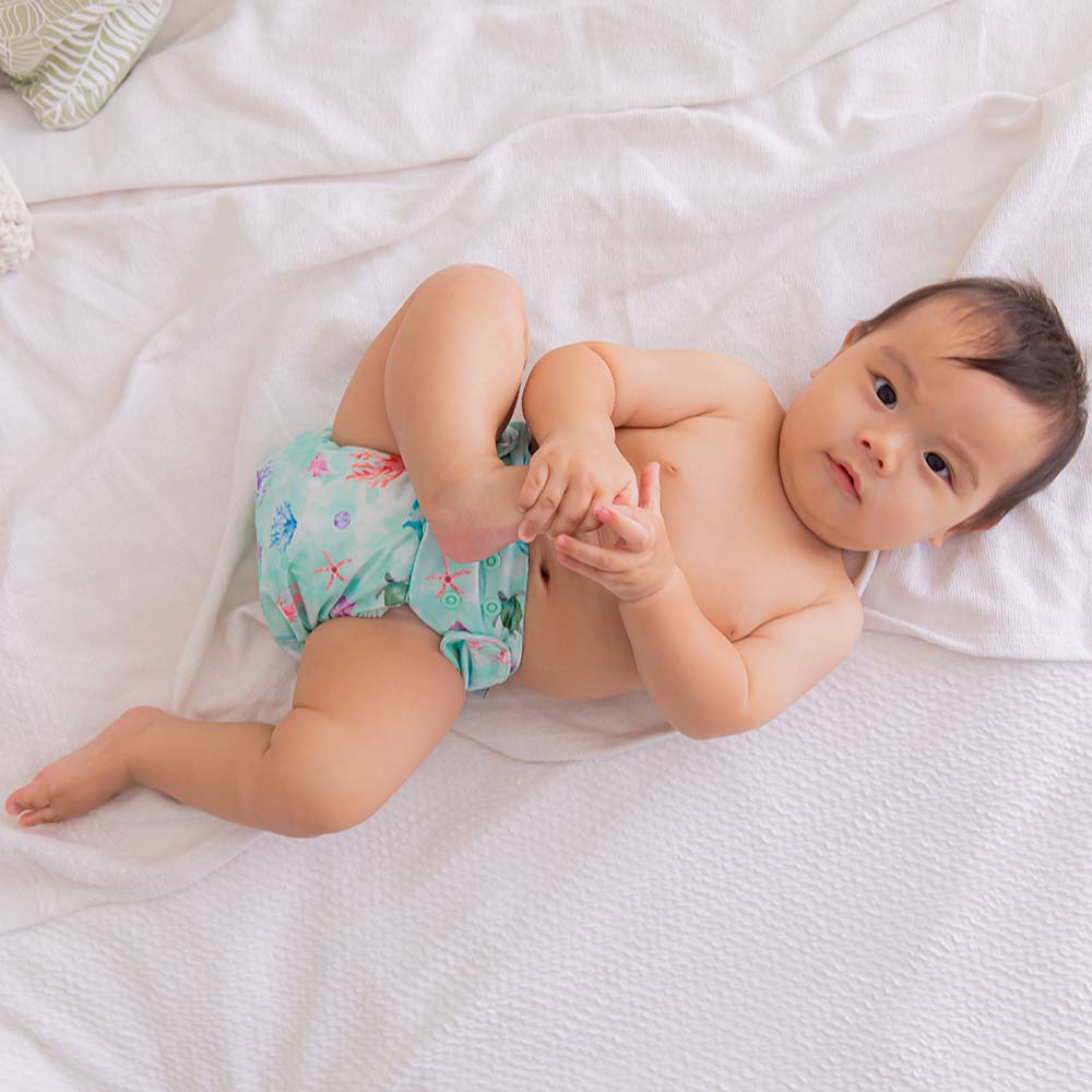 Baby looking at camera holding his foot and laying on the bed, Wearing oceanic cloth nappy