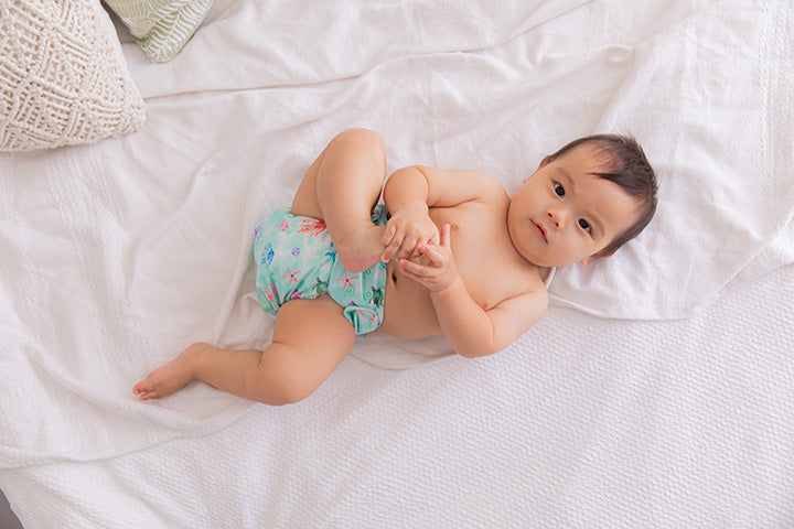 Baby on a white bed holding his foot wearing just an ocean themed cloth nappy