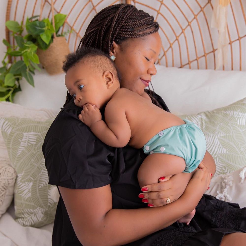 Black mother with eyes closed cuddling young baby close to her. Baby wearing sage green cloth nappy.