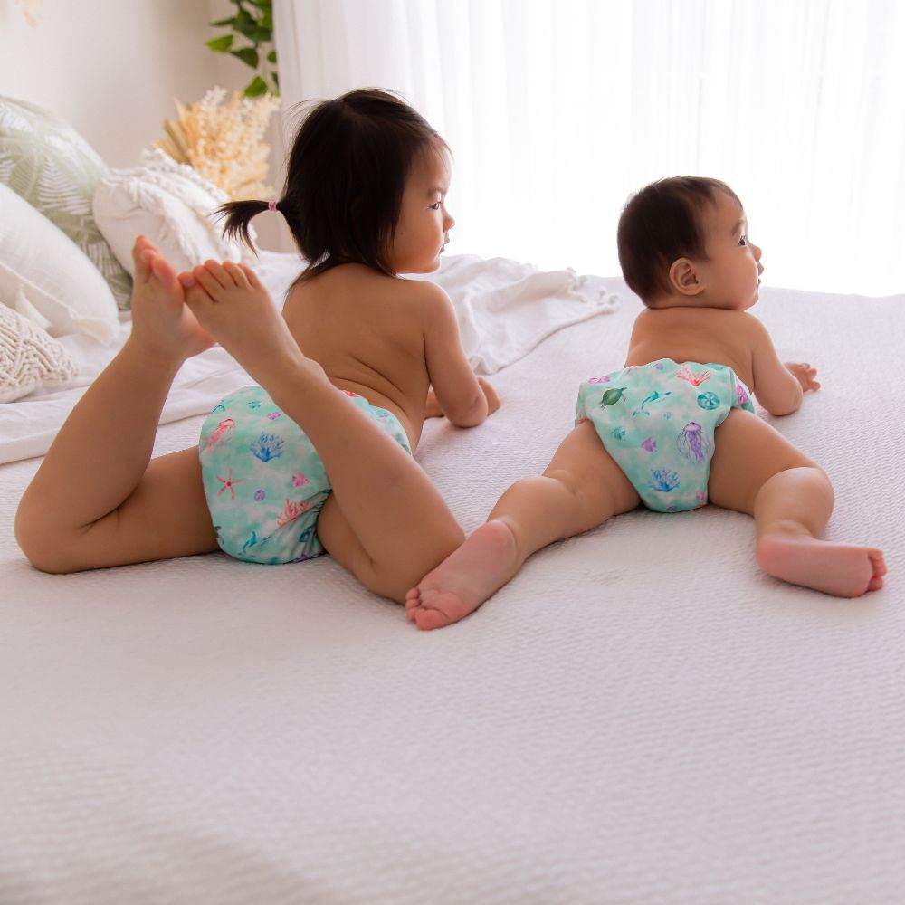 Asian siblings laying on their stomachs on a bed looking to the side, wearing matching oceanic reusable nappies.