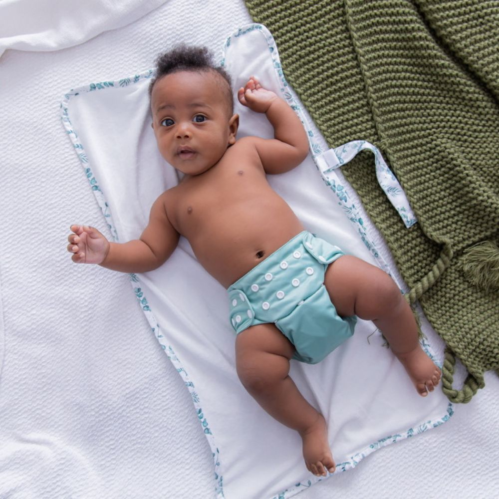 Image shows young black baby looking at camera laying on a portable change mat wearing sage green reusable nappy.