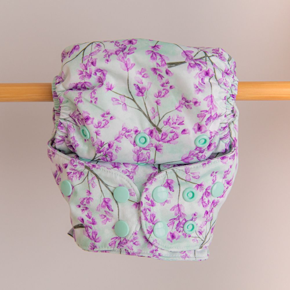 Purple and green jacaranda print reusable nappy attached upside down to a wooden bar