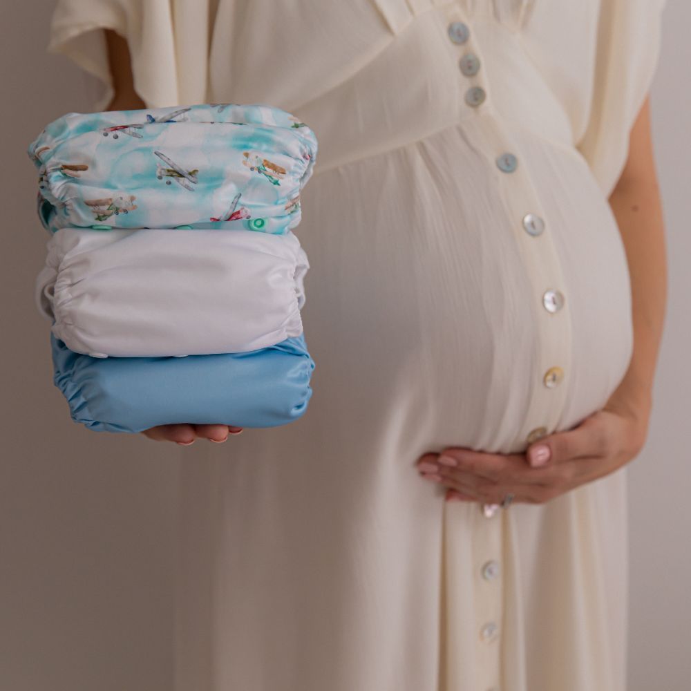 Close up of an expectant mother's baby bump, she cradles her bump whilst holding a stack of blue, white and aeroplane cloth nappy prints with her other hand